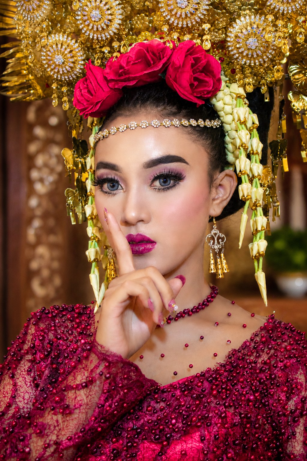 Close-up beauty portrait: wajah, makeup, mahkota emas dengan mawar merah, dan gesture jari telunjuk di bibir